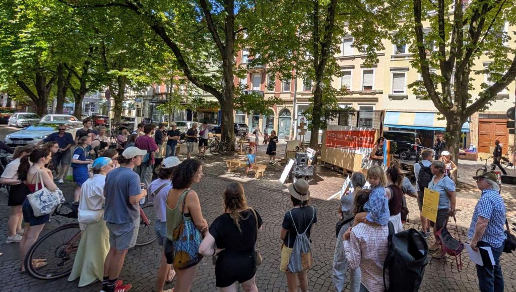 Menschen stehen auf dem Werderplatz um eine Rednerin. In der Mitte steht ein Plakat mit der Aufschrift "Eigentum verpflichtet!"
