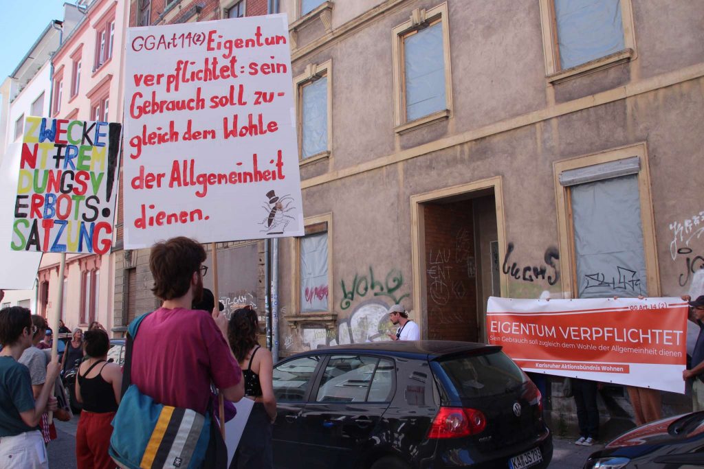 Teilnehmer einer Demonstration stehen vor Häusern, deren Fenster mit Folie abgedeckt sind. Auf den Transparenten der Demonstranten liest man: "Eigentum verpflichtet. Sein Gebrauch soll zugleich dem Wohle der Allgemeinheit dienen." und "Zweckentfremdungsverbotssatzung".